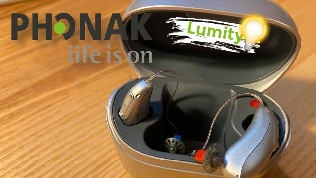 Zwei Phonak Lumity Hörgeräte in einer offenen Ladestation, auf Holztisch fotografiert