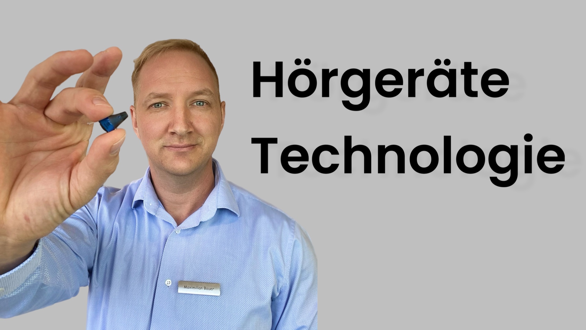 Maximilian Bauer mit kleinem Hörgerät in der Hand. Im Hintergrund der Schriftzug "Hörgeräte Technologie"
