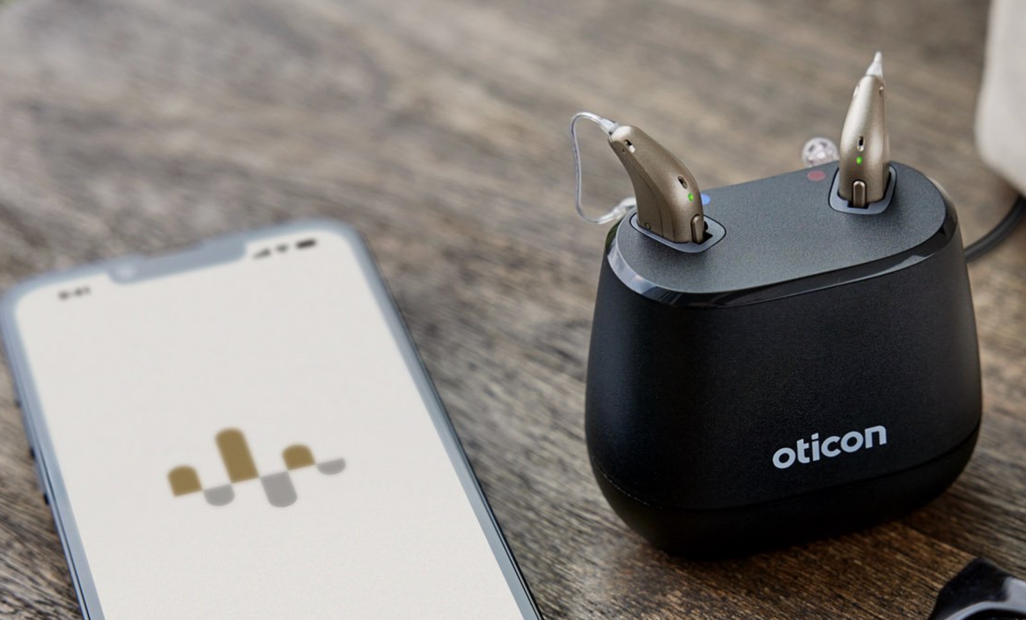 Oticon Intent wird in Ladestation geladen. Daneben Liegt ein Handy mit Oticon App